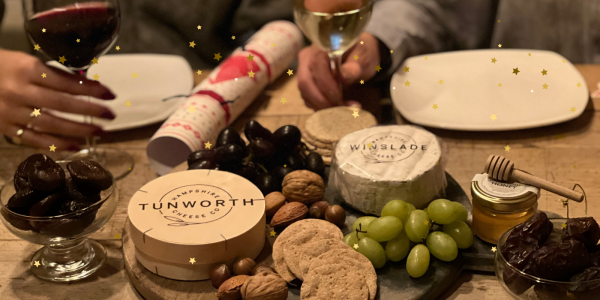 Image shows Tunworth and Winslade Cheese on a Slate Cheeseboard with Nuts, Crackers, Green and Red Grapes, with Two Cheese Plates, A Glass of Red and White Wine and a Christmas Cracker